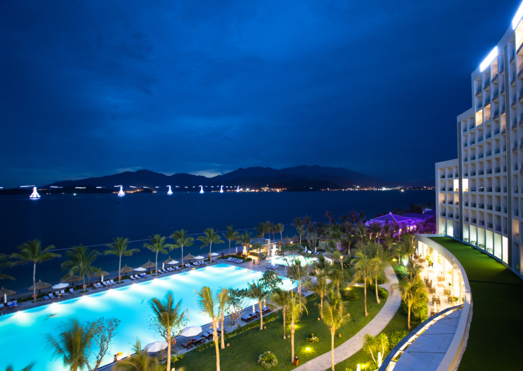 Começo da noite em uma paisagem que mostra o mar ao fundo. Na frente, uma grande piscina iluminada, com espreguiçadeiras, árvores, uma piscina melhor e parte de um grande prédio na lateral da imagem.