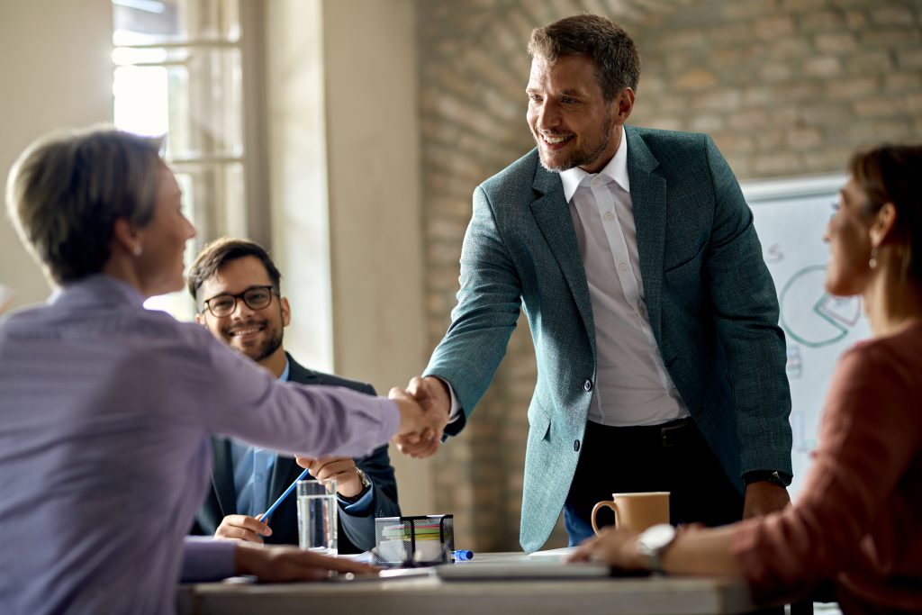 Reunião de negócios, com três homens e uma mulher usando roupas com estilo profissional. Dois homens se cumprimentam com um aperto de mãos.