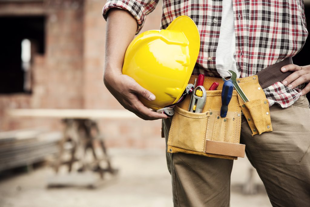 Cintura de um mestre de obras, com um cinto para guardar pequenas ferramentas de trabalho. Na mão, ele segura um capacete de segurança e, ao fundo da imagem, pode-se ver uma obra em andamento.