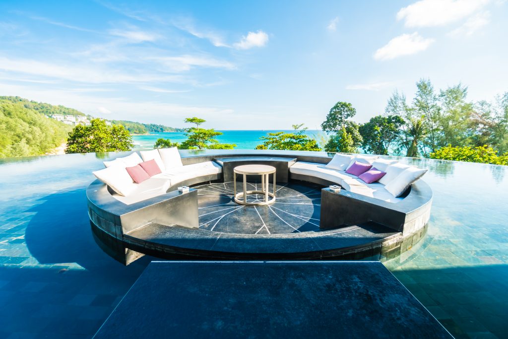 Piscina com borda infinita e o mar atrás, além de elementos naturais. No centro da piscina, há uma plataforma redonda e seca, com sofás e uma mesa de centro.